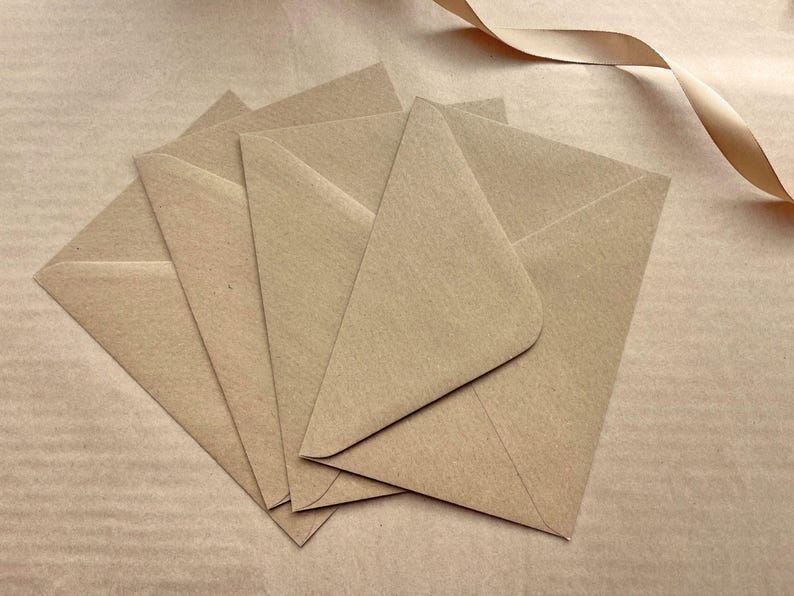 May include: A collection of five kraft paper envelopes fanned out, with a light brown ribbon in the upper right corner. The envelopes are arranged to show their triangular shape and the texture of the paper.