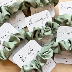 Peut inclure: Un ensemble de chouchous en satin vert avec des cartes blanches attach&eacute;es. Les cartes portent diff&eacute;rents titres, notamment "Bride Tribe", "Maid of Honor", "Flower Girl" et "Bridesmaid".