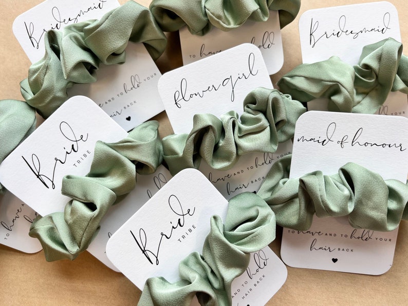 Peut inclure: Un ensemble de chouchous en satin vert avec des cartes blanches attach&eacute;es. Les cartes portent diff&eacute;rents titres, notamment "Bride Tribe", "Maid of Honor", "Flower Girl" et "Bridesmaid".
