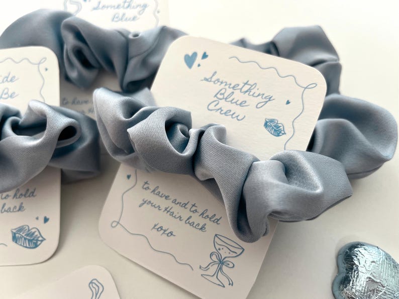 Puede incluir: Coletero de sat&eacute;n azul claro con tarjetas. Las tarjetas dicen "Something Blue Crew" y "to have and to hold your Hair back xoxo", con ilustraciones de coraz&oacute;n, labios y copa de champ&aacute;n.