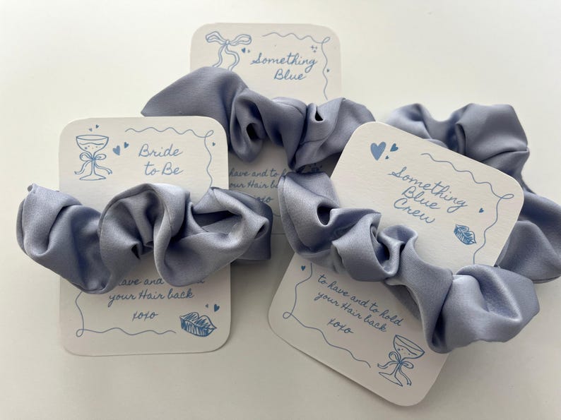 Puede incluir: Coleteros de sat&eacute;n azul claro con tarjetas blancas. Las tarjetas tienen ilustraciones azules y texto, incluyendo "Bride to Be" y "Something Blue Crew". Las tarjetas tambi&eacute;n dicen "to have and to hold your hair back".
