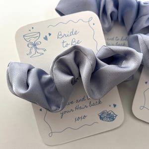 Puede incluir: Coleteros de sat&eacute;n azul claro con tarjetas "Bride to Be". Las tarjetas tienen una ilustraci&oacute;n de copa de champ&aacute;n y el texto "to have and to hold your hair back xoxo". Ideales para despedidas de soltera.
