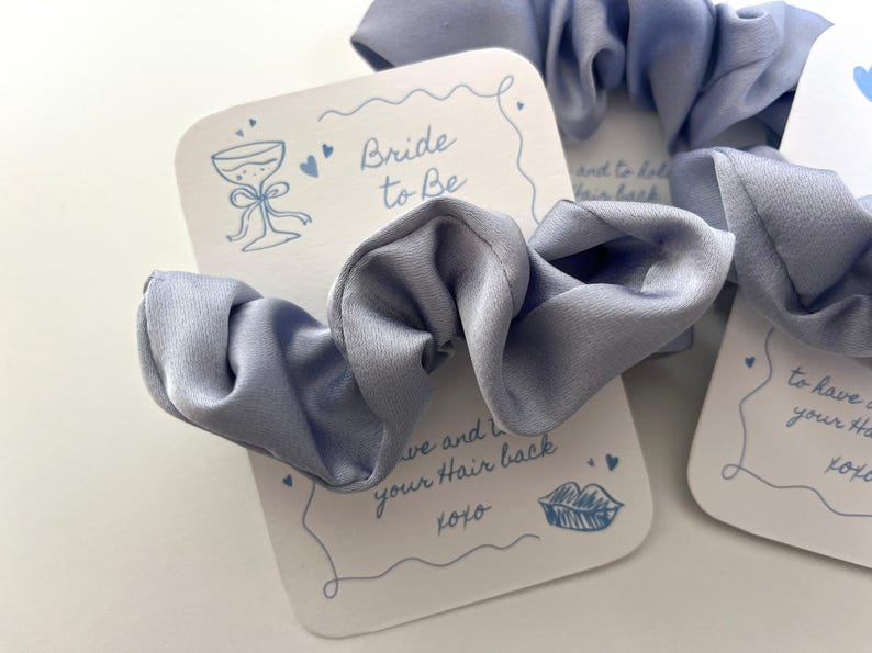 Puede incluir: Coleteros de sat&eacute;n azul claro con tarjetas "Bride to Be". Las tarjetas tienen una ilustraci&oacute;n de copa de champ&aacute;n y el texto "to have and to hold your hair back xoxo". Ideales para despedidas de soltera.