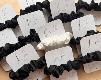 May include: Black satin scrunchies with white cards that read "I do" and "Crew". One white scrunchie is labeled "Bride". A wedding favor idea.