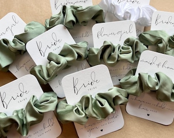 May include: Set of green satin scrunchies with white cards that say "Bride Tribe", "Maid of Honor", "Flower Girl", and "To have and to hold your hair back".