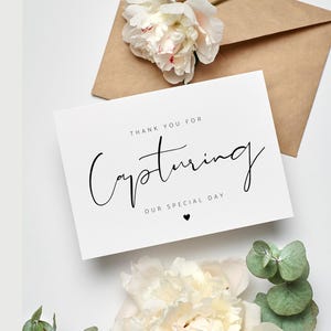 May include: A white card with the text "THANK YOU FOR CAPTURING OUR SPECIAL DAY" in black script. The card is next to a brown envelope and white flowers with green foliage.