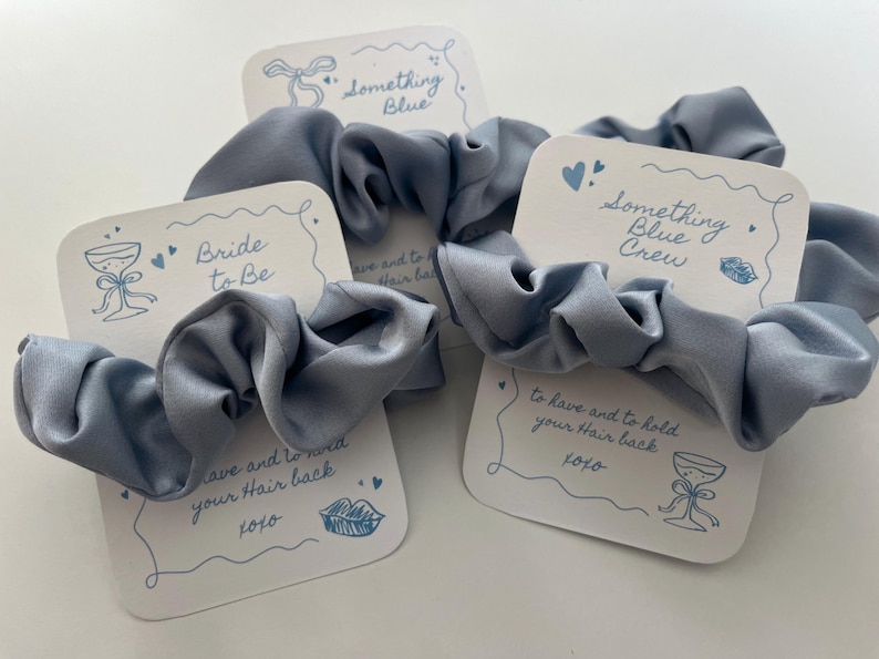 Puede incluir: Tres coleteros azul claro con tarjetas blancas. Las tarjetas tienen las frases "Bride to Be" y "Something Blue Crew" en escritura azul. Cada tarjeta tiene un dibujo de una copa de champ&aacute;n y el texto "to have and to hold your hair back xoxo."