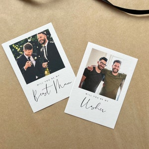 May include: Two white cards featuring photographs of men, with the text "Will you be my Best Man" and "Will you be my Usher" in cursive script. The cards are on a brown surface.