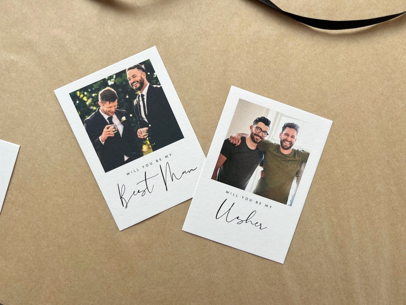 May include: Two white cards featuring photographs of men, with the text "Will you be my Best Man" and "Will you be my Usher" in cursive script. The cards are on a brown surface.