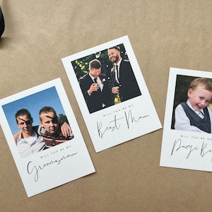 May include: Three white cards with photos and text on a brown surface. The cards read "Will you be my Groomsman", "Will you be my Best Man", and "Will you be our Page Boy". A black ribbon is in the upper left corner.