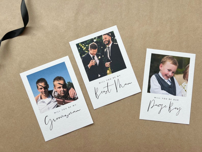 May include: Three white cards with photos and text on a brown surface. The cards read "Will you be my Groomsman", "Will you be my Best Man", and "Will you be our Page Boy". A black ribbon is in the upper left corner.