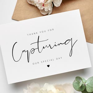May include: A white card with the text "Thank you for Capturing Our Special Day" in elegant black script. A small black heart is below the text. The card is next to a brown envelope and white flowers.