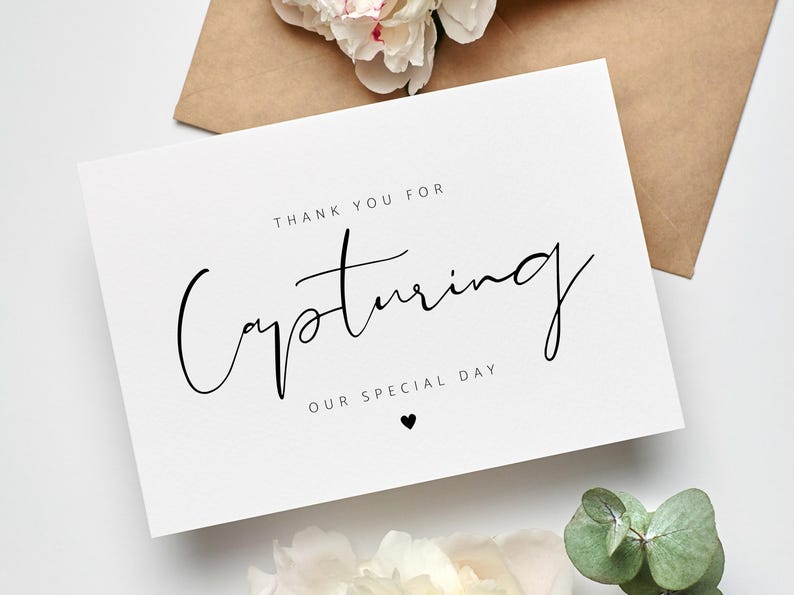 May include: A white card with the text "Thank you for Capturing Our Special Day" in elegant black script. A small black heart is below the text. The card is next to a brown envelope and white flowers.