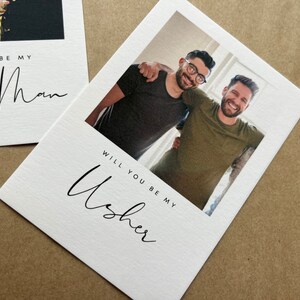 May include: White cards with black script reading "Will you be my Usher" and "Will you be my Best Man." One card features a photo of two men. The cards are on a brown surface.