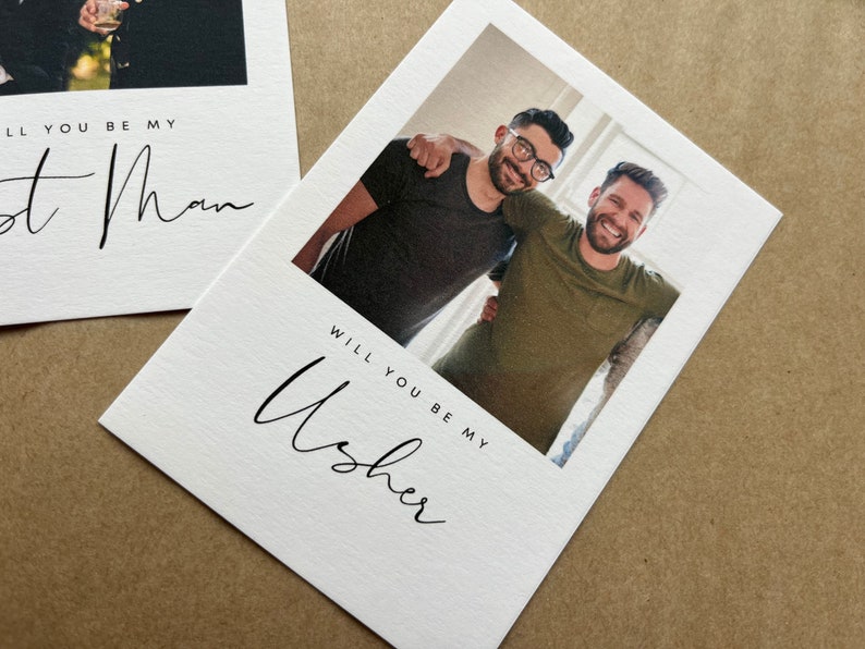 May include: White cards with black script reading "Will you be my Usher" and "Will you be my Best Man." One card features a photo of two men. The cards are on a brown surface.