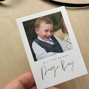 May include: A white card featuring a photograph of a smiling young boy in a grey waistcoat and white shirt. The card reads "WILL YOU BE OUR PAGE BOY" in black script.