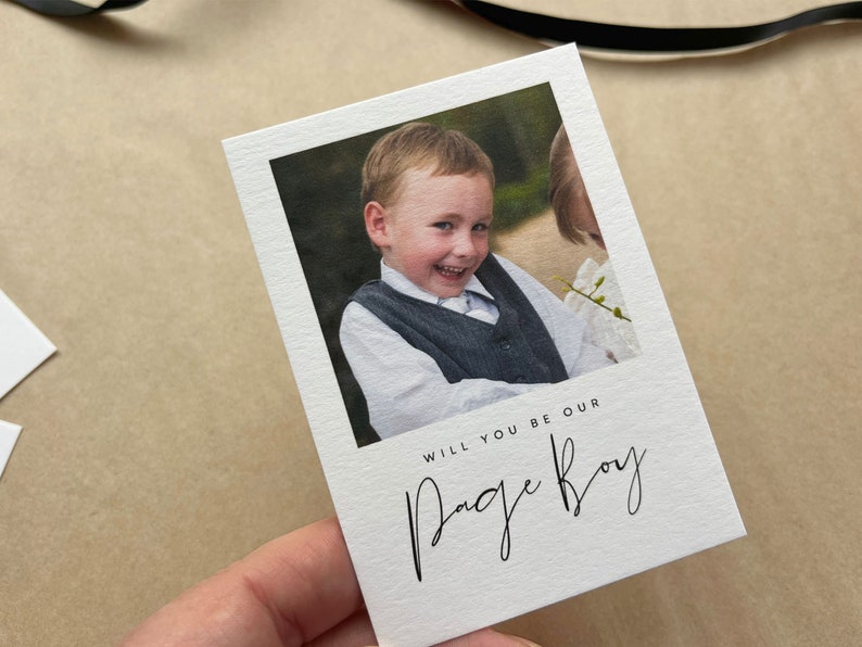 May include: A white card featuring a photograph of a smiling young boy in a grey waistcoat and white shirt. The card reads "WILL YOU BE OUR PAGE BOY" in black script.
