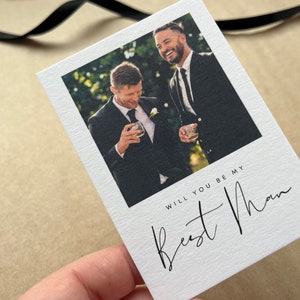 May include: White card featuring a photograph of two men in suits, holding glasses. The card has the text "WILL YOU BE MY Best Man" in black script. A black ribbon is visible.