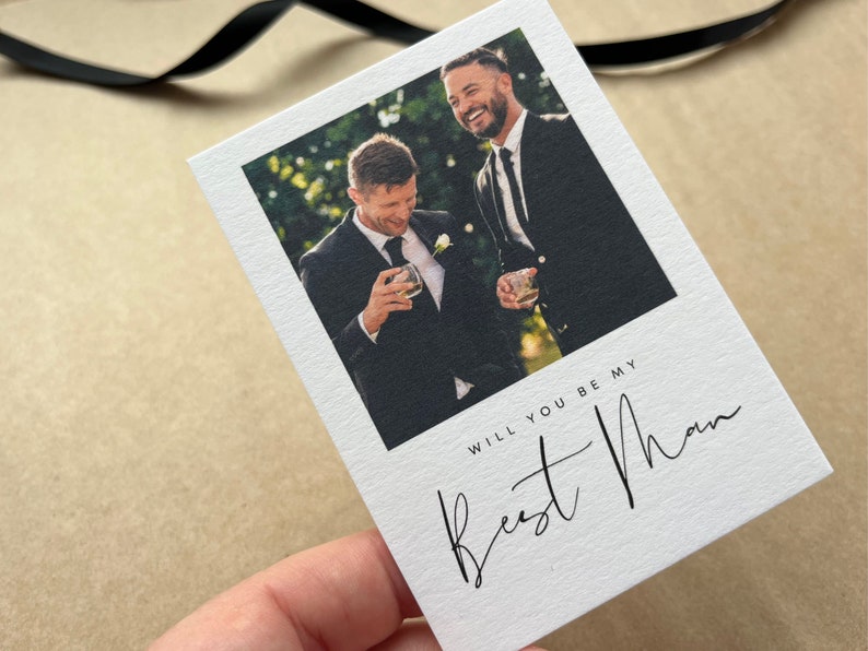 May include: White card featuring a photograph of two men in suits, holding glasses. The card has the text "WILL YOU BE MY Best Man" in black script. A black ribbon is visible.