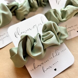 Peut inclure: Une carte blanche avec le texte "flower girl" et "to have and to hold your hair back" en &eacute;criture noire. Un chouchou vert sauge est plac&eacute; sur la carte.