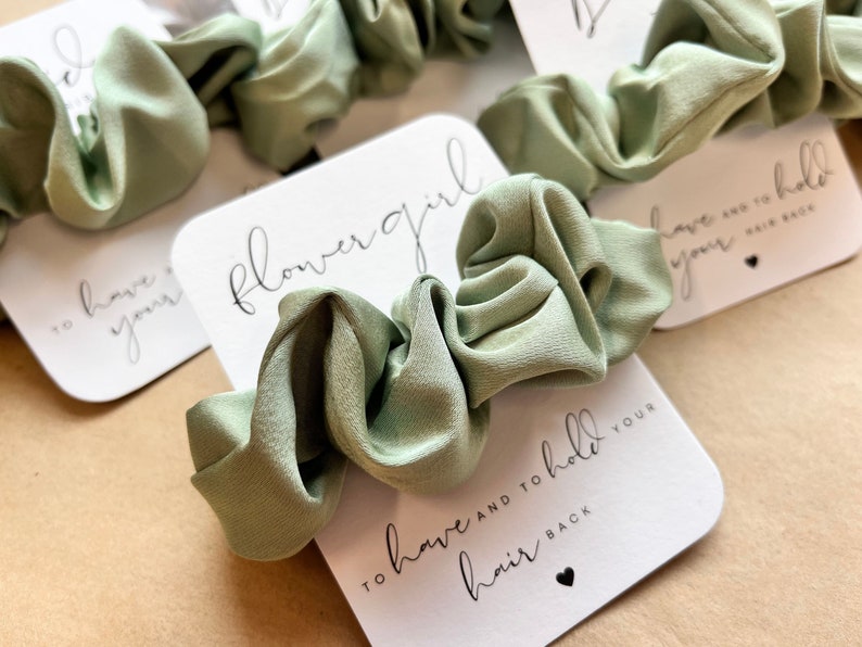 Peut inclure: Une carte blanche avec le texte "flower girl" et "to have and to hold your hair back" en &eacute;criture noire. Un chouchou vert sauge est plac&eacute; sur la carte.