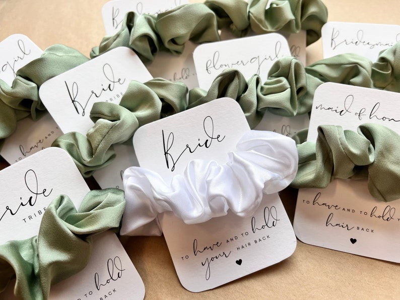 Peut inclure: Un ensemble de chouchous pour cheveux avec des cartes qui disent "Bride Tribe", "Bride", "Maid of Honor", "Bridesmaid" et "Flower Girl". Les chouchous sont en satin vert et blanc.