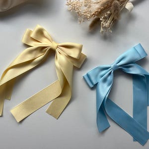 May include: Two satin hair bows, one in a pale yellow and the other in light blue, are shown on a white surface. Each bow features long, flowing ribbons. Dried flowers and a piece of fabric are in the background.