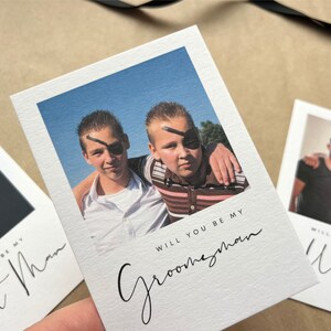 May include: White invitation cards with photos and text. One card says "Will you be my Groomsman" with a photo of two boys wearing eye patches. Other cards say "Best Man" and "Usher".