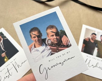 May include: White invitation cards with photos and text. One card says "Will you be my Groomsman" with a photo of two boys. Another says "Will you be my Best Man" with a photo of a man. A third says "Will you be my Usher".