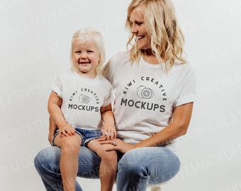 May include: A woman and a child wearing white t-shirts with the text "Kiwi Creative Mockups" and a graphic of a kiwi fruit. The woman is sitting on a white stool and the child is sitting on her lap. Both are wearing blue jeans.