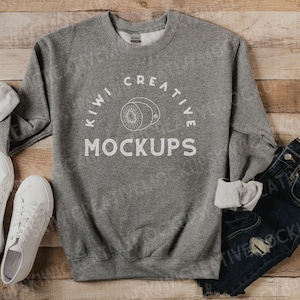 May include: A gray crewneck sweatshirt with the text "KIWI CREATIVE MOCKUPS" and a kiwi fruit graphic. The sweatshirt is paired with white sneakers and dark blue jeans on a wooden surface.