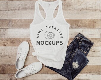 May include: White tank top with a black graphic of two kiwi fruit and the text "Kiwi Creative Mockups". The tank top is laying on a wooden surface with a pair of white sneakers and blue jeans.