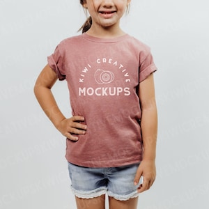 May include: A young person wearing a dusty rose-colored t-shirt with rolled-up sleeves and denim shorts. The shirt features the text "KIWI CREATIVE MOCKUPS" in a circular design. The shorts have a white lace trim.