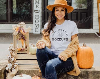 Op de afbeelding: Een vrouw draagt een wit T-shirt met de tekst "Kiwi Creative Mockups" in zwarte letters, een bruine cardigan, een blauwe jeans en een bruine hoed. Ze zit op een houten trede met pompoenen en herfstdecoraties om haar heen.