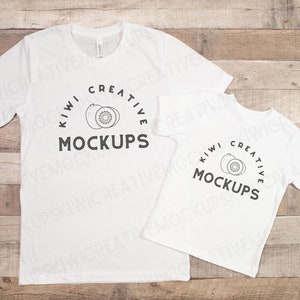 Può includere: Due magliette bianche con una grafica in bianco e nero di un kiwi e il testo "Kiwi Creative Mockups". Le magliette sono disposte su una superficie di legno.