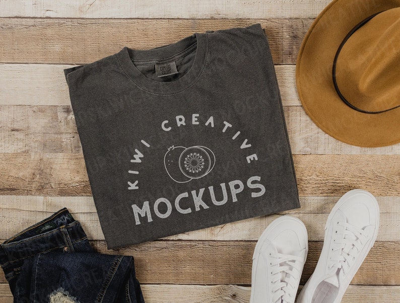 Op de afbeelding: Een grijs T-shirt met de tekst "KIWI CREATIVE MOCKUPS" erop gedrukt. Het shirt ligt op een houten oppervlak met een bruine hoed, een blauwe spijkerbroek en witte sneakers.