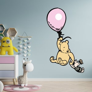 Puede incluir: Una calcoman&iacute;a de pared de Winnie the Pooh con Winnie the Pooh y Piglet agarrados a un globo rosa. La calcoman&iacute;a est&aacute; en una pared azul claro con una c&oacute;moda y otros juguetes para ni&ntilde;os en el fondo.