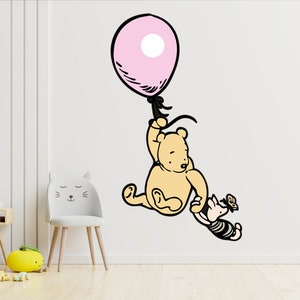 Puede incluir: Una calcoman&iacute;a de pared de Winnie de Pooh y Piglet que muestra a Winnie de Pooh sosteniendo un globo rosa y Piglet agarr&aacute;ndose del brazo de Winnie de Pooh. La calcoman&iacute;a est&aacute; en una pared blanca.