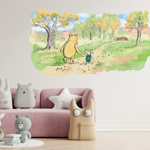 May include: A Winnie the Pooh and Piglet wall decal featuring the two characters walking hand-in-hand along a forest path. The decal is set against a white wall with a pink sofa and plush toys in the foreground.