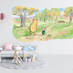 May include: A Winnie the Pooh and Piglet wall decal featuring a watercolour illustration of the two characters walking along a path in a forest. The decal is on a white wall in a nursery with a rocking horse, a doll, and a cloud-shaped pillow.