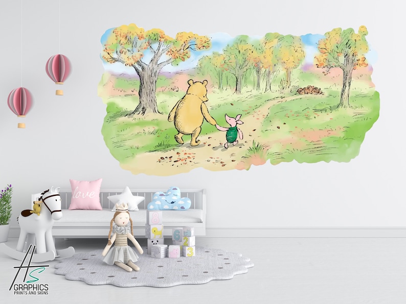 May include: A Winnie the Pooh and Piglet wall decal featuring a watercolour illustration of the two characters walking along a path in a forest. The decal is on a white wall in a nursery with a rocking horse, a doll, and a cloud-shaped pillow.