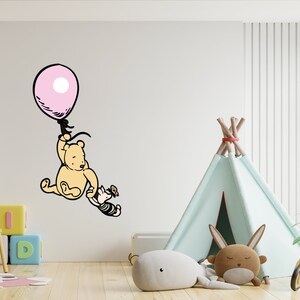 Puede incluir: Una calcoman&iacute;a de pared de Winnie de Pooh y Piglet con Winnie de Pooh sosteniendo un globo rosa y Piglet alcanz&aacute;ndolo. La calcoman&iacute;a est&aacute; en una pared blanca en una guarder&iacute;a con una tienda de campa&ntilde;a tipi, un juguete de madera y una planta.
