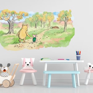 May include: A Winnie the Pooh and Piglet wall decal featuring a watercolour illustration of the characters walking down a path in a forest. The decal is displayed on a white wall in a child's room with a white table, blue bench, and pink chair.