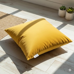 Puede incluir: Un cojín cuadrado de terciopelo amarillo mostaza descansa sobre un suelo de madera clara. El cojín está en primer plano, con una pequeña etiqueta en la esquina inferior derecha. La luz del sol proyecta sombras sobre el suelo y el cojín. Dos pequeñas plantas en maceta están en el fondo.