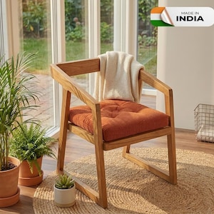 Puede incluir: Sillón de madera con un cojín de pana naranja quemado y una manta color crema sobre el respaldo. La silla está sobre una alfombra de yute, con plantas en macetas cerca. La imagen incluye el texto "MADE IN INDIA".
