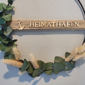 Pu&ograve; includere: Una ghirlanda in metallo nero con un nastro di juta e lettere in legno che compongono la parola "HEIMATHAFEN". La ghirlanda &egrave; decorata con foglie di eucalipto ed erba della pampa secca.