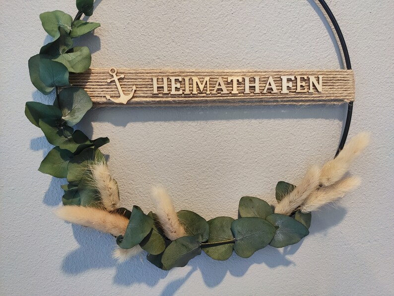 Pu&ograve; includere: Una ghirlanda in metallo nero con un nastro di juta e lettere in legno che compongono la parola "HEIMATHAFEN". La ghirlanda &egrave; decorata con foglie di eucalipto ed erba della pampa secca.