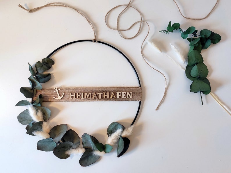 Pu&ograve; includere: Una ghirlanda in metallo nero con uno striscione in juta che recita "HEIMATHAFEN" e una piccola ancora in legno. La ghirlanda &egrave; decorata con foglie di eucalipto e fiori secchi bianchi e soffici.