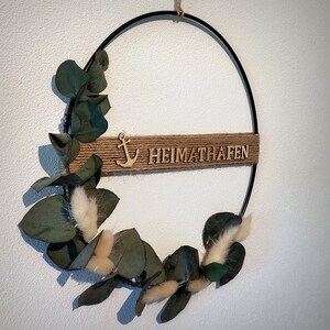 Pu&ograve; includere: Una ghirlanda di metallo nero con uno striscione di juta che recita "Heimathafen" e un'ancora di legno. La ghirlanda &egrave; decorata con foglie di eucalipto e fiori bianchi e soffici.