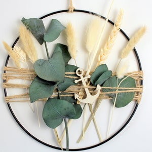 Könnte beinhalten: Ein schwarzer Metallreifenkranz, der mit getrockneten Naturmaterialien wie Weizen, Pampasgras und Eukalyptusblättern verziert ist. Ein Anker aus Holz befindet sich in der Mitte des Kranzes.